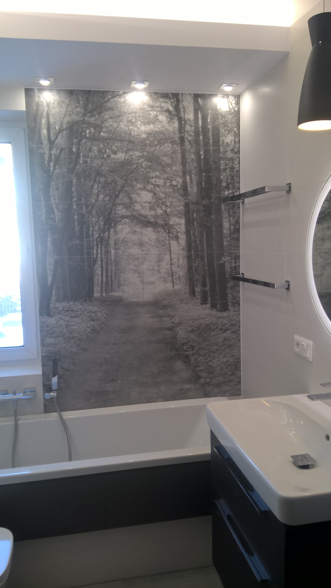 Nowoczesna łazienka z wanną, umywalką z szafką i ścianą wyłożoną płytkami z motywem czarno-białego lasu, oświetlona punktowymi lampami sufitowymi i wiszącą lampą nad lustrem.
