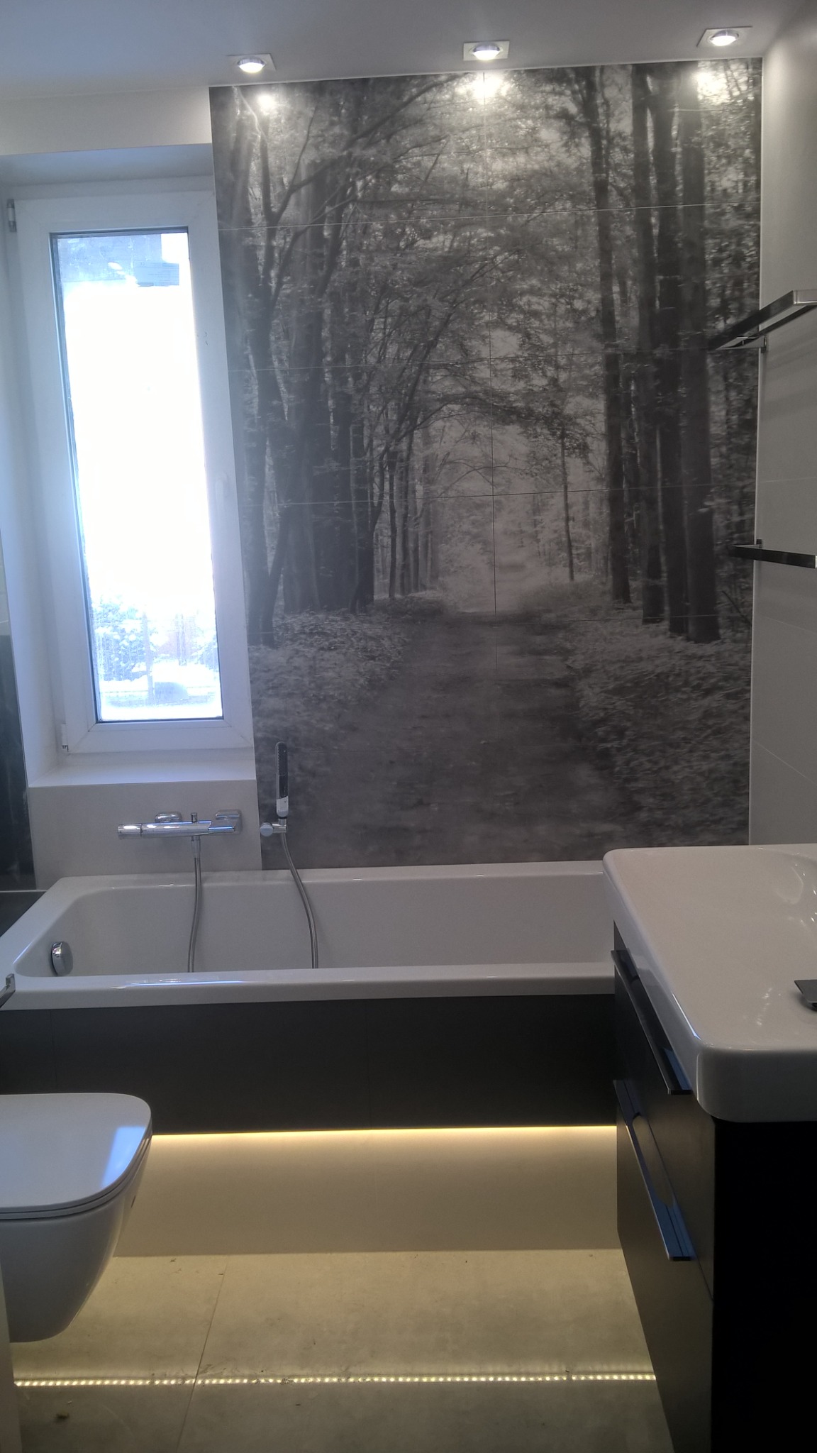 Nowoczesna łazienka w Tarnowskich Górach z wanną, umywalką i toaletą. Na ścianie położono płytki z motywem leśnej ścieżki w odcieniach szarości. Podświetlenie LED pod wanną i szafką umywalkową.