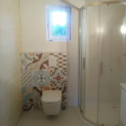 Wykończona łazienka z kabiną prysznicową, toaletą podwieszaną i ścianą z dekoracyjnych płytek wzorzystych, widok na okno i drewnianą boazerię.