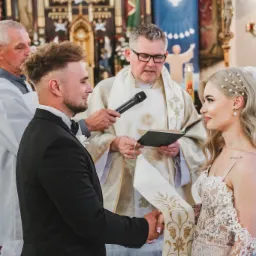 Para młoda trzymająca się za ręce podczas ceremonii ślubnej w kościele, w tle ksiądz z mikrofonem i ministrant