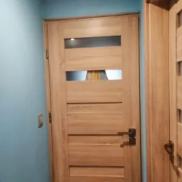 Dębowa wewnętrzna drzwi z poziomymi panelami i trzema przeszkleniami, zamontowana w pomieszczeniu z błękitnymi ścianami, widoczny fragment ościeżnicy drugiej pary drzwi.