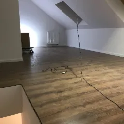 Poddasze po remoncie z nowymi panelami podłogowymi w odcieniach brązu, widoczne kable elektryczne i grzejnik, częściowo oświetlone naturalnym światłem z okna dachowego.