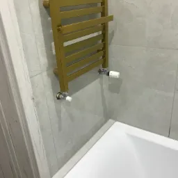 Złoty grzejnik drabinkowy zamontowany na szarej, marmurkowej ścianie w łazience, widoczna biała wanna i fragment dekoracyjnej, drewnopodobnej obudowy.