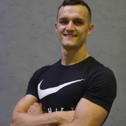 Uśmiechnięty mężczyzna z zielonymi oczami, w czarnej koszulce z logo Nike, stoi na tle szarej ściany z rękami skrzyżowanymi na piersiach.