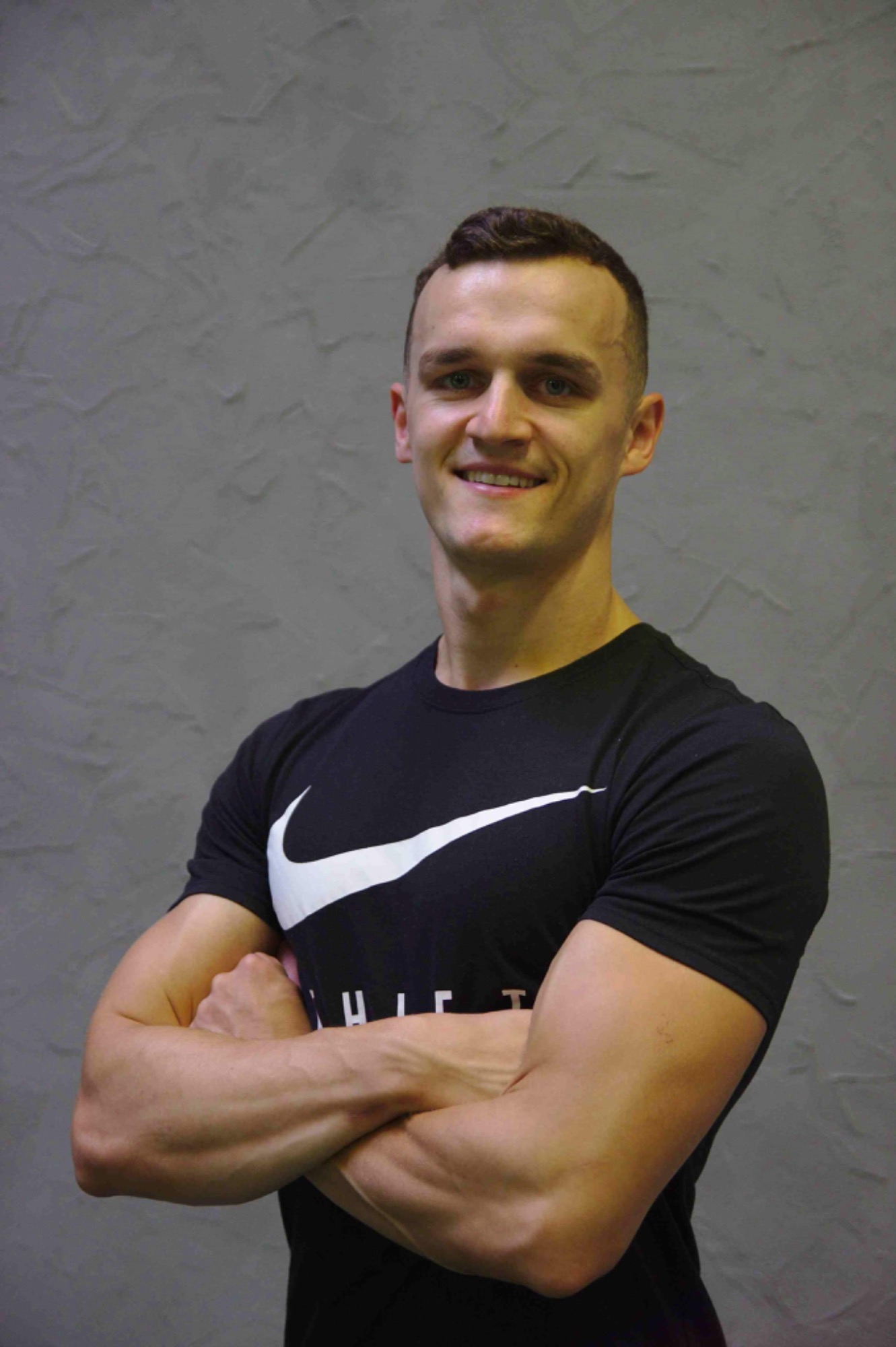 Uśmiechnięty mężczyzna z zielonymi oczami, w czarnej koszulce z logo Nike, stoi na tle szarej ściany z rękami skrzyżowanymi na piersiach.