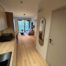 MARMAL - Jasne wnętrze mieszkania z drewnianą podłogą, widok z kuchni na salon z sofą i balkonem. Na ścianie owalne lustro. Minimalistyczny design.