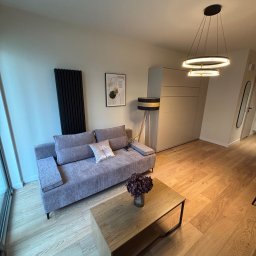 MARMAL - Nowoczesny salon z szarą sofą, drewnianym stolikiem kawowym i czarnym grzejnikiem panelowym. Minimalistyczny design, ciepłe oświetlenie, drewniana podłoga.