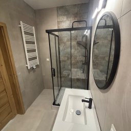 MARMAL - Nowoczesna łazienka po remoncie: kabina prysznicowa z czarnymi profilami, umywalka z szafką, okrągłe lustro i grzejnik drabinkowy na ścianie w odcieniach szarości.