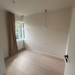 Jasne, puste wnętrze pokoju po remoncie: parkiet, kremowe ściany, okno z zasłoną i minimalistyczne lampy wiszące. Widok zza otwartych drzwi.