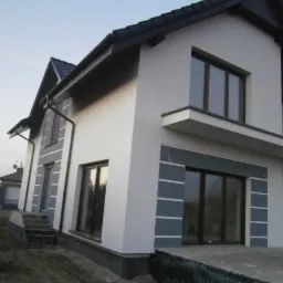 Dwukondygnacyjny dom z jasną elewacją i grafitowymi akcentami wokół okien. Dach z ciemnej dachówki. Widok z zewnątrz, w tle inne budynki.