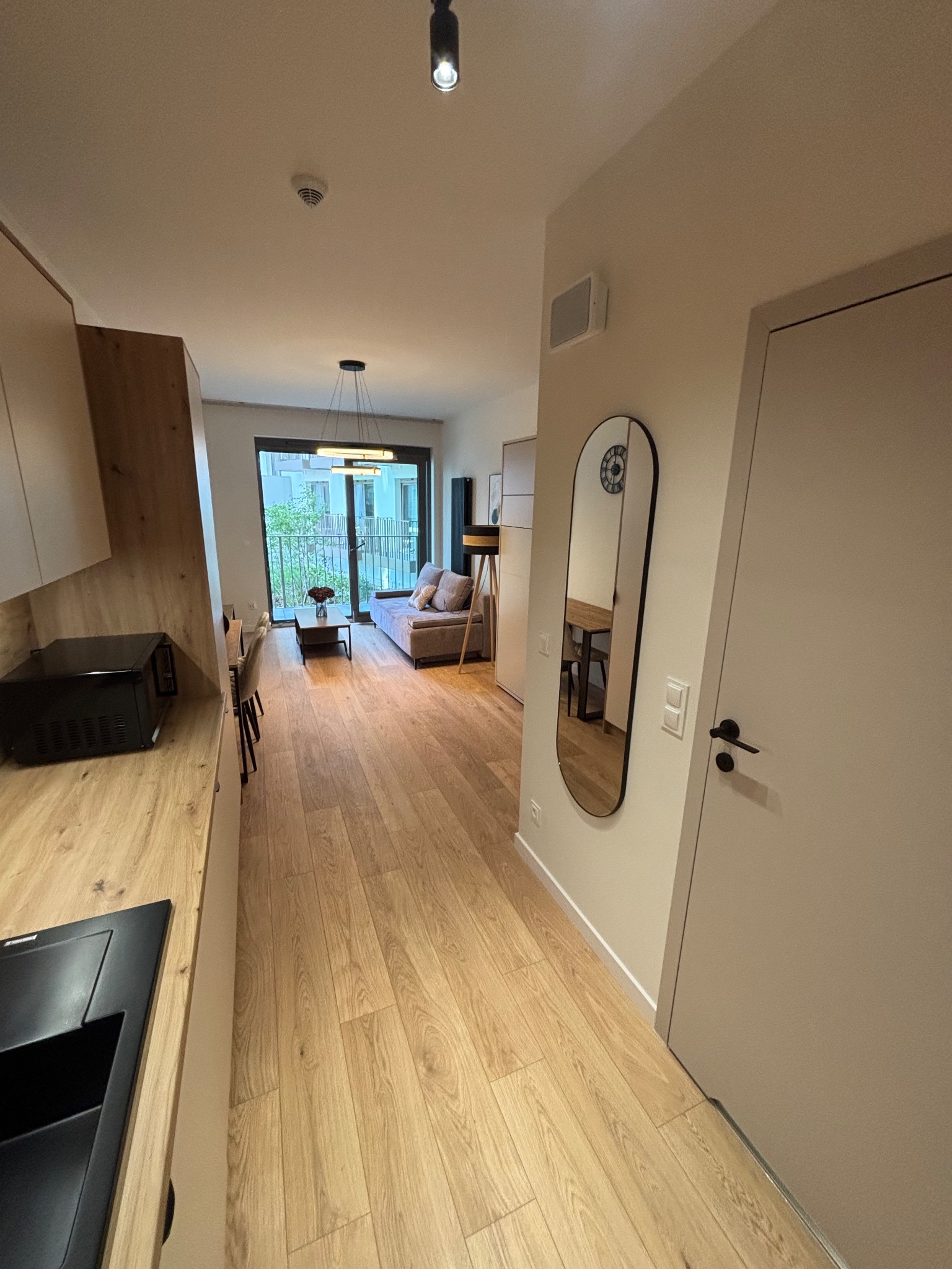 Jasne wnętrze mieszkania z drewnianą podłogą, widok z kuchni na salon z sofą i balkonem. Na ścianie owalne lustro. Minimalistyczny design.