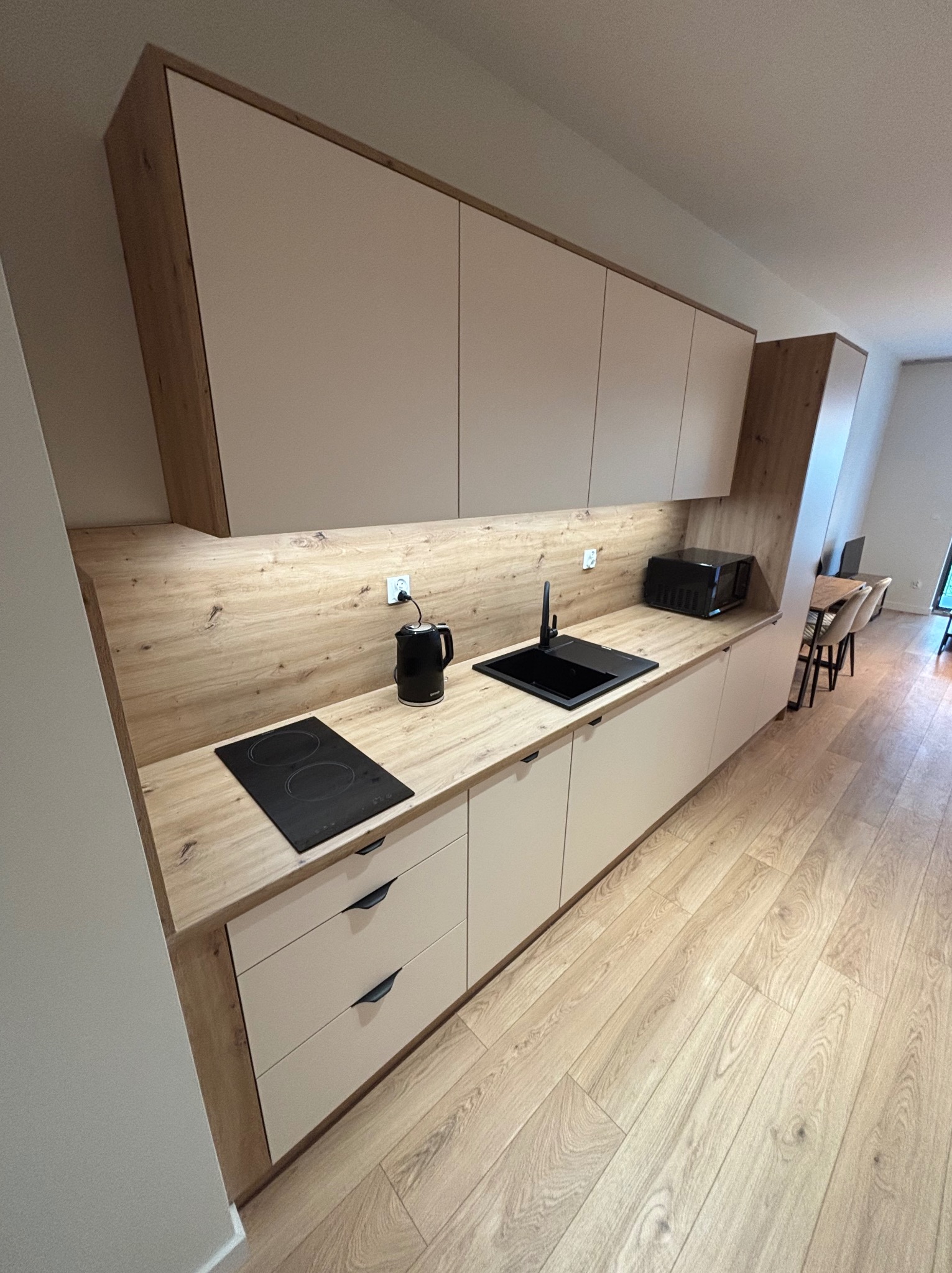 Nowoczesna kuchnia z drewnianymi akcentami: beżowe szafki, czarny zlew i płyta indukcyjna, drewniany blat i podłoga. Minimalistyczny design z oświetleniem podszafkowym.