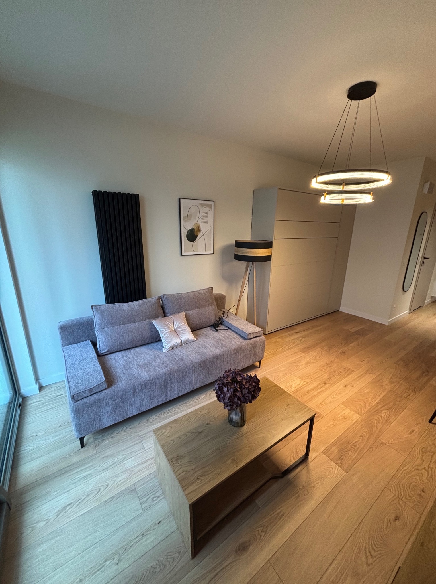 Nowoczesny salon z szarą sofą, drewnianym stolikiem kawowym i czarnym grzejnikiem panelowym. Minimalistyczny design, ciepłe oświetlenie, drewniana podłoga.