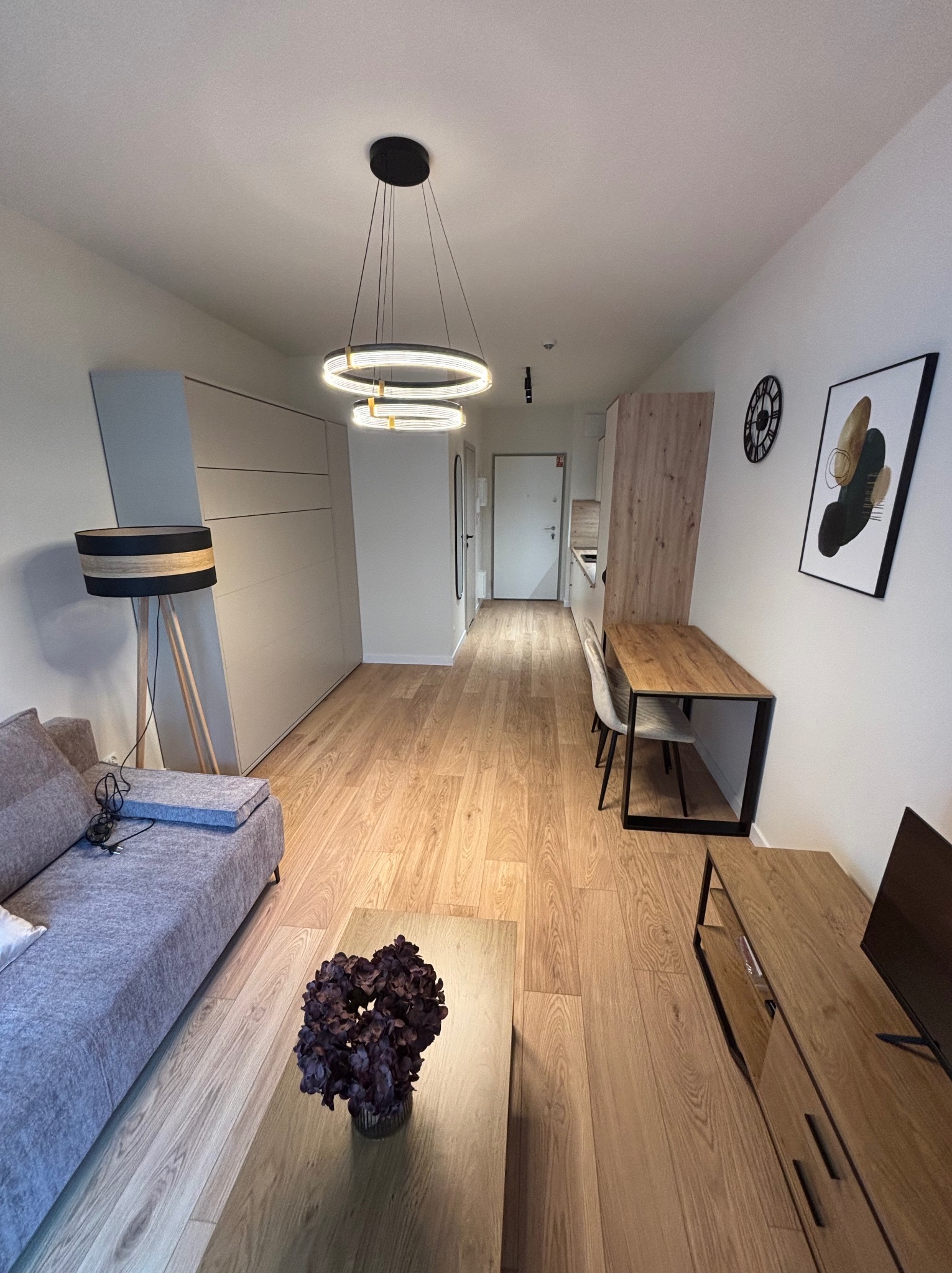 Nowoczesne wnętrze mieszkania z drewnianą podłogą, szarą sofą, okrągłym żyrandolem i minimalistycznym wystrojem. Widok na stół z krzesłami i dekoracje ścienne.