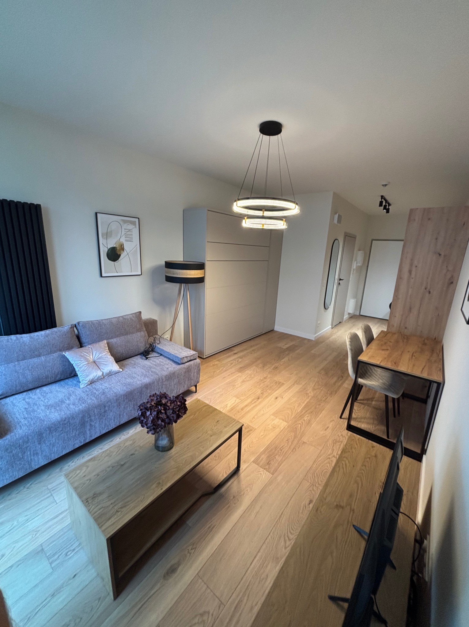 Jasne wnętrze mieszkania z drewnianą podłogą, szarą sofą, stolikiem kawowym i lampą. Minimalistyczny design z nowoczesnym oświetleniem i dekoracjami.