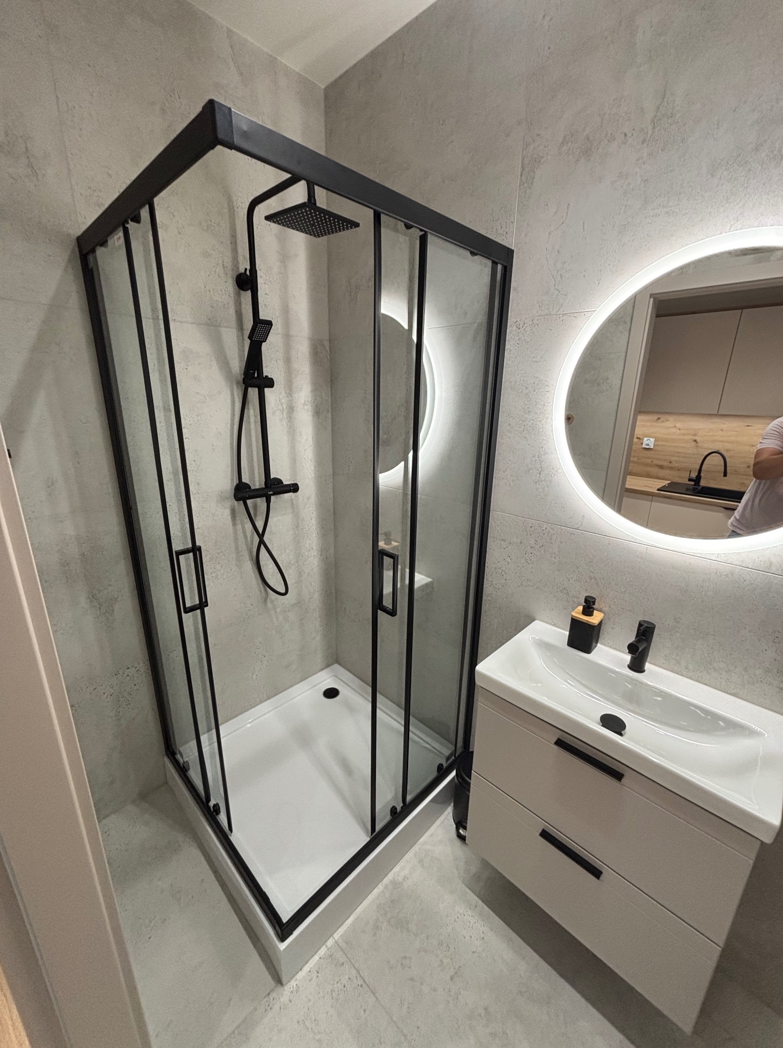 Nowoczesna łazienka z kabiną prysznicową typu walk-in z czarnymi profilami, białą umywalką z szafką i okrągłym lustrem z podświetleniem LED. Minimalistyczny design.