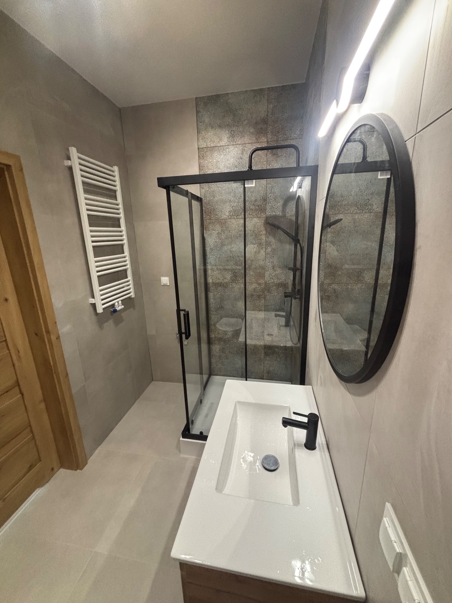 Nowoczesna łazienka po remoncie: kabina prysznicowa z czarnymi profilami, umywalka z szafką, okrągłe lustro i grzejnik drabinkowy na ścianie w odcieniach szarości.