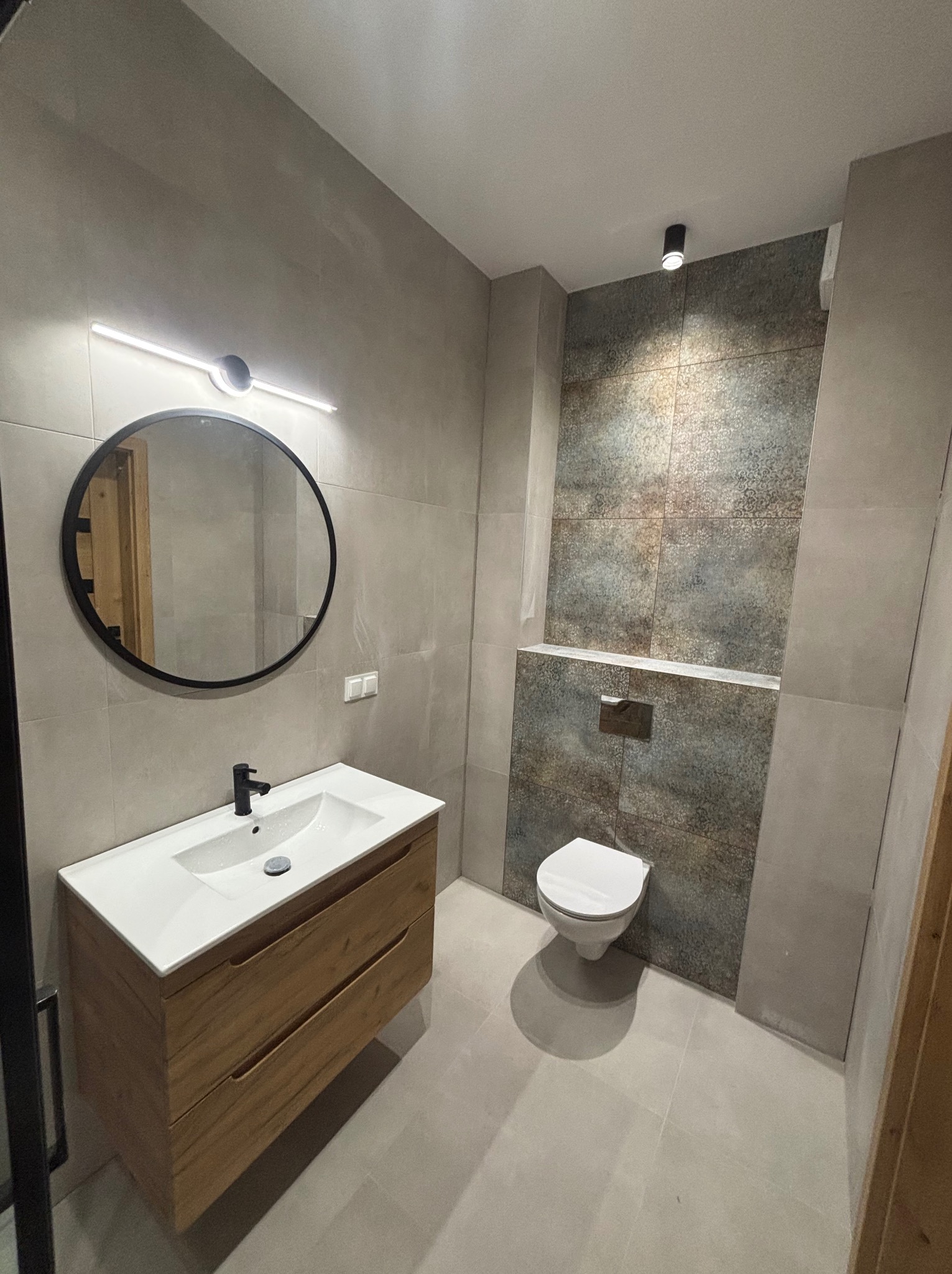 Wykończona łazienka w szarościach z drewnianą szafką pod umywalką i okrągłym lustrem z czarną ramą. Na ścianie dekoracyjne płytki za toaletą.