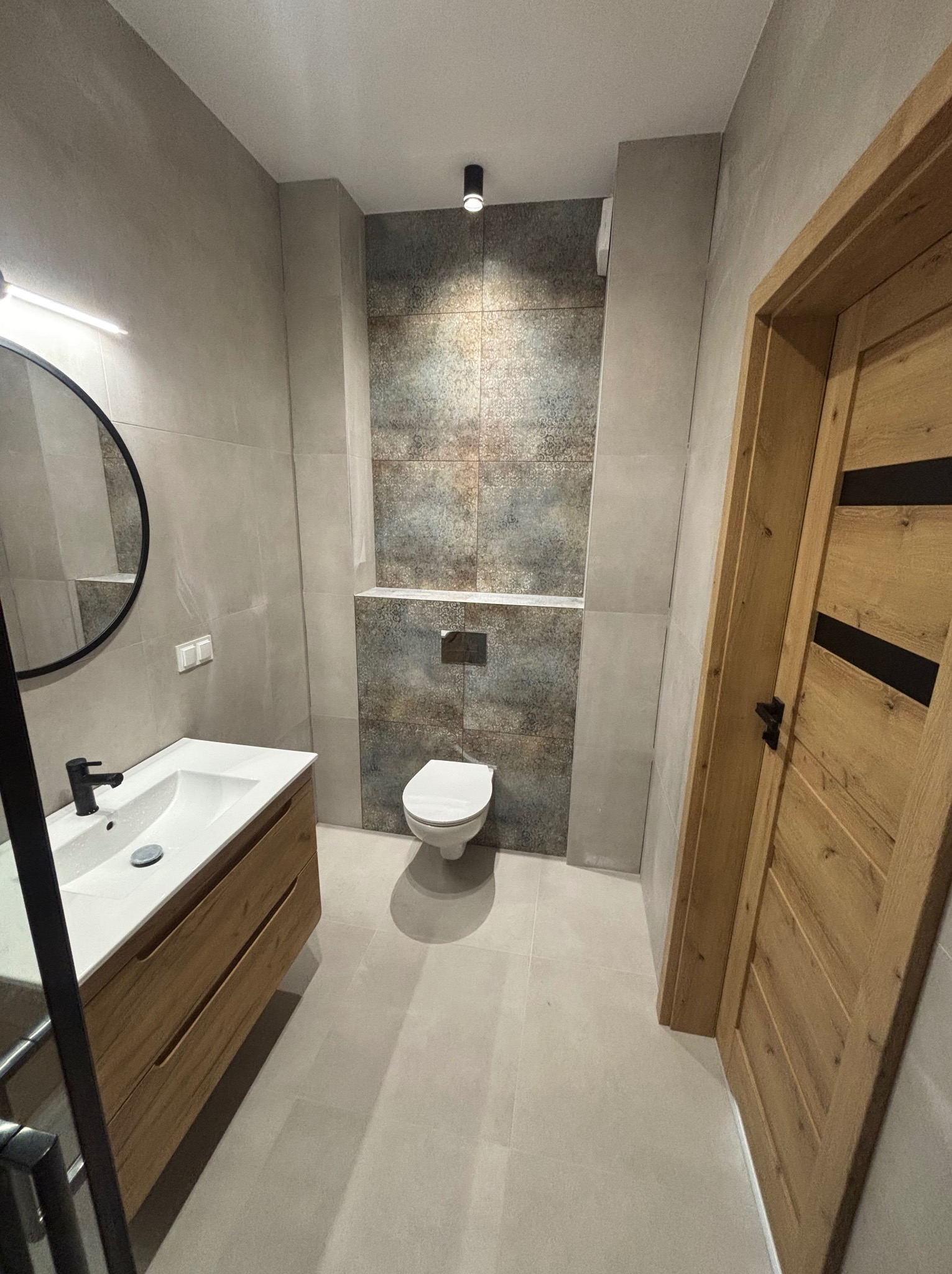 Nowoczesna łazienka z umywalką na drewnianej szafce, okrągłym lustrem i toaletą w zabudowie. Szare płytki i drewniane drzwi tworzą spójny design.