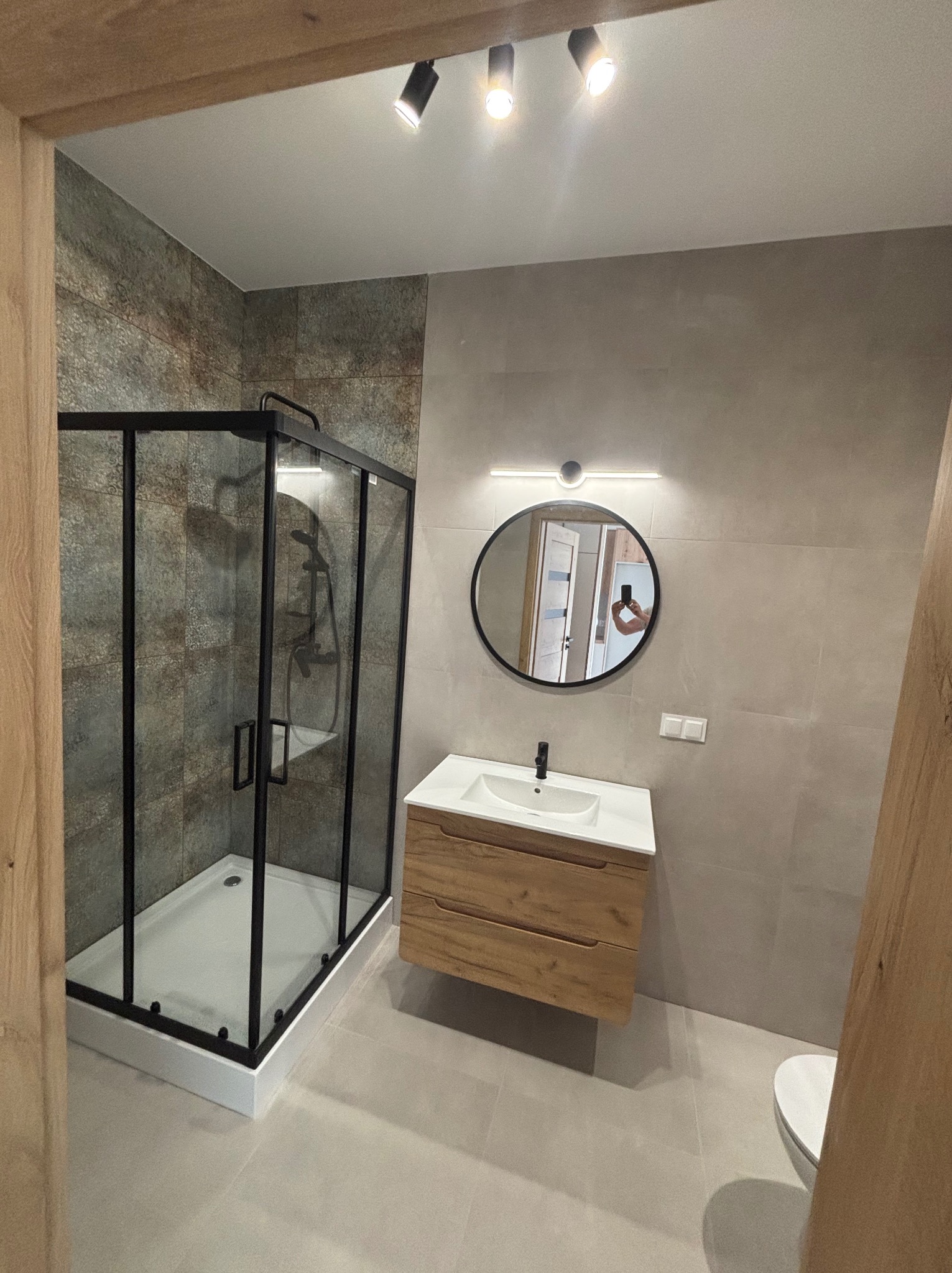 Nowoczesna łazienka z kabiną prysznicową w czarnej ramie, drewnianą szafką pod umywalką i okrągłym lustrem z podświetleniem. Stonowana kolorystyka płytek.