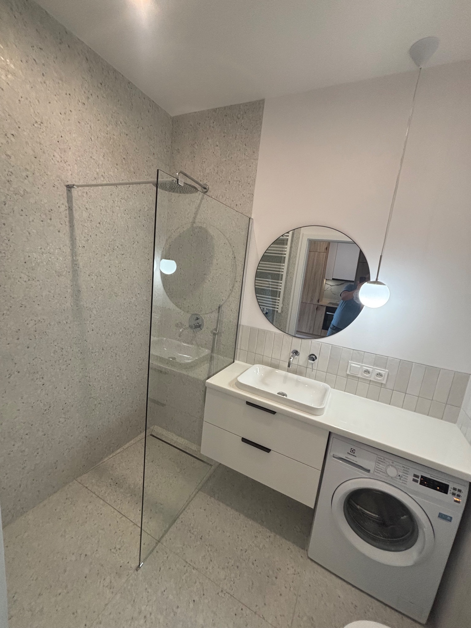 Nowoczesna łazienka z kabiną prysznicową typu walk-in, umywalką nablatową z szafką, pralką i okrągłym lustrem. Ściany i podłoga wykończone płytkami w odcieniach szarości.