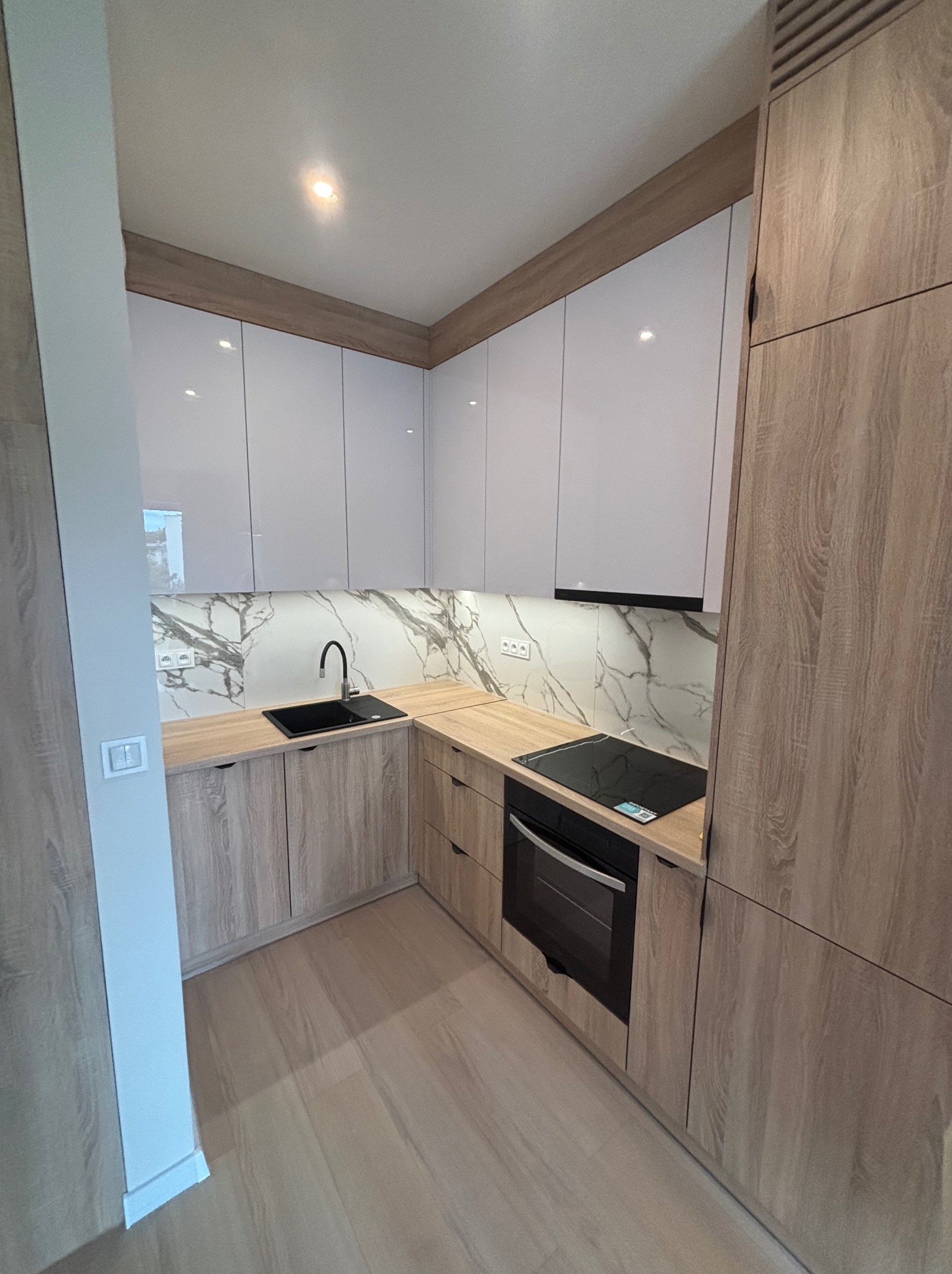 Nowoczesna kuchnia w kształcie litery L z białymi szafkami górnymi i drewnopodobnymi dolnymi, czarnym zlewem i płytą, marmurowym backsplashiem i jasną podłogą.