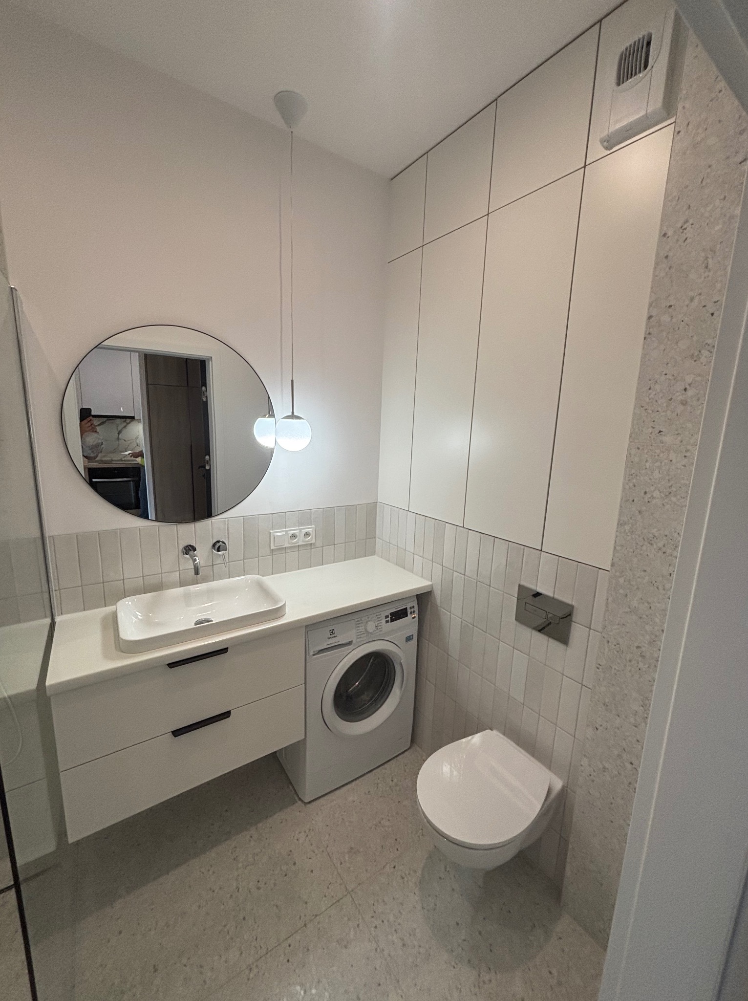 Nowoczesna łazienka w bieli i szarości: umywalka z szafką, pralka, toaleta podwieszana, okrągłe lustro i płytki imitujące cegłę. Minimalistyczny design.