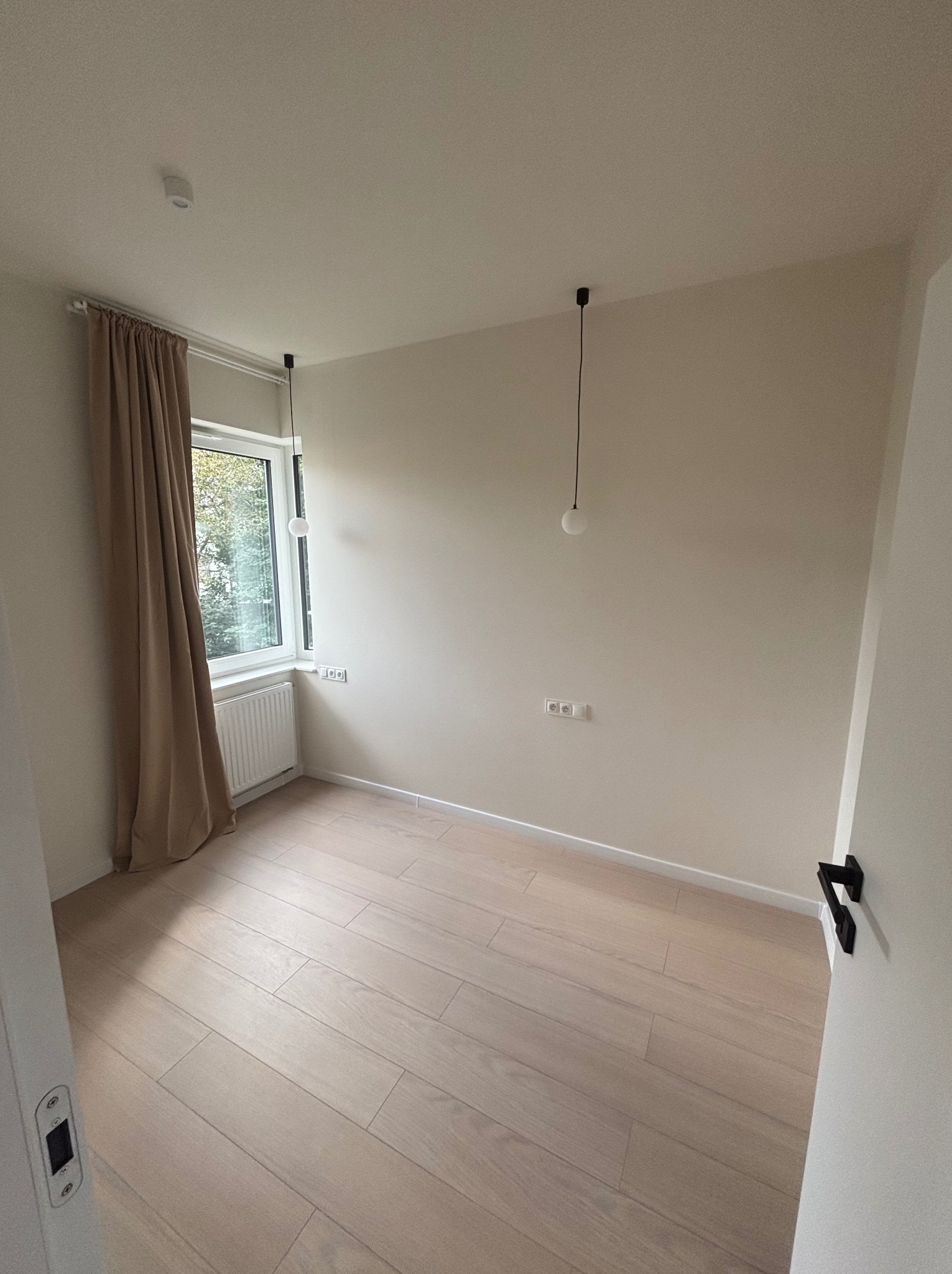 Jasne, puste wnętrze pokoju po remoncie: parkiet, kremowe ściany, okno z zasłoną i minimalistyczne lampy wiszące. Widok zza otwartych drzwi.