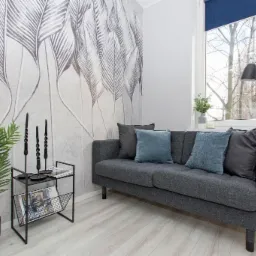 Jasny salon z szarą sofą, wzorzystą tapetą w liście, drewnianą podłogą, czarnymi świecami na metalowym stoliku i rośliną doniczkową.