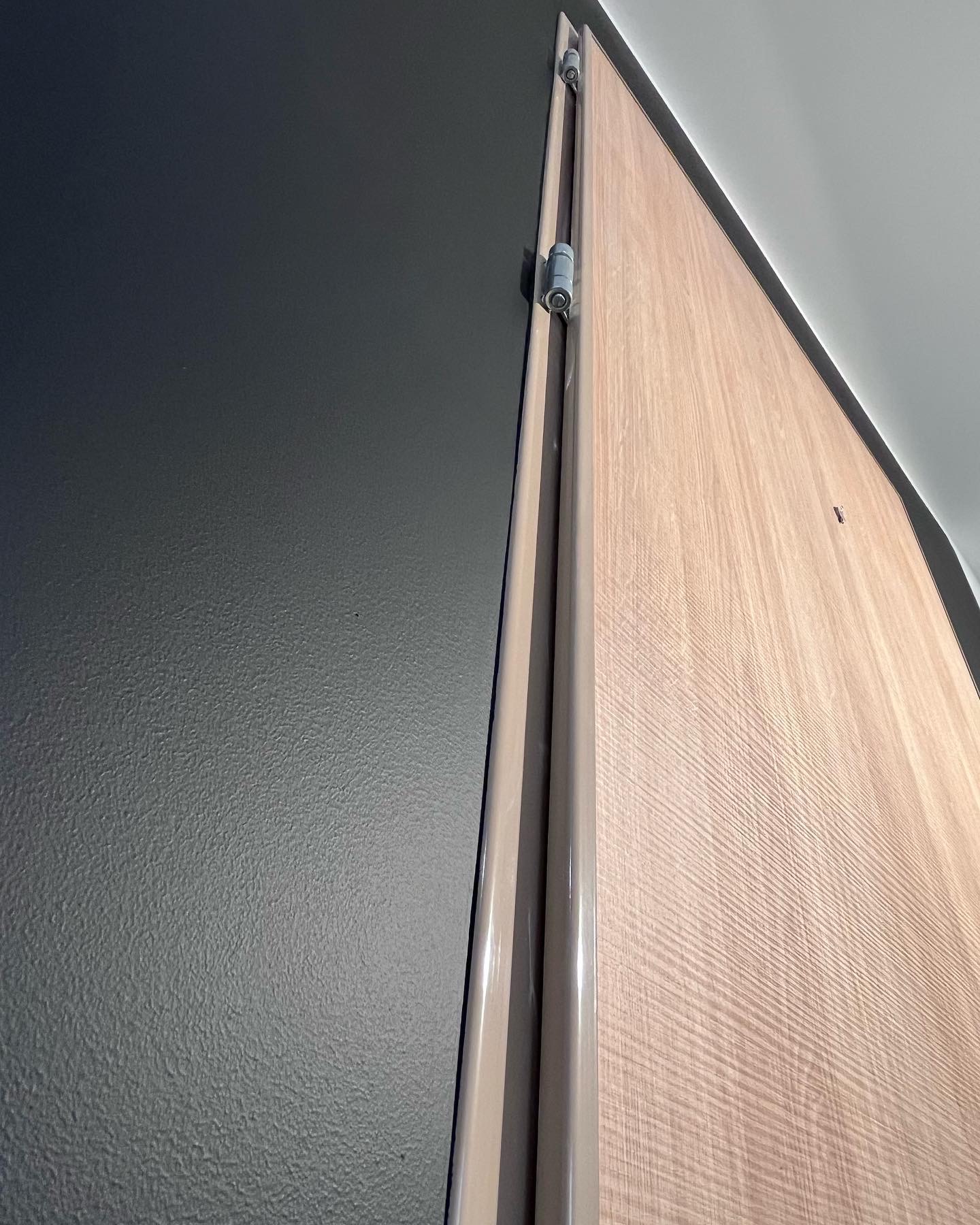 Fragment ściany pomalowanej na ciemny szary kolor, sąsiadującej z jasną, drewnianą ścianą z widocznymi listwami wykończeniowymi i ukrytymi zawiasami.