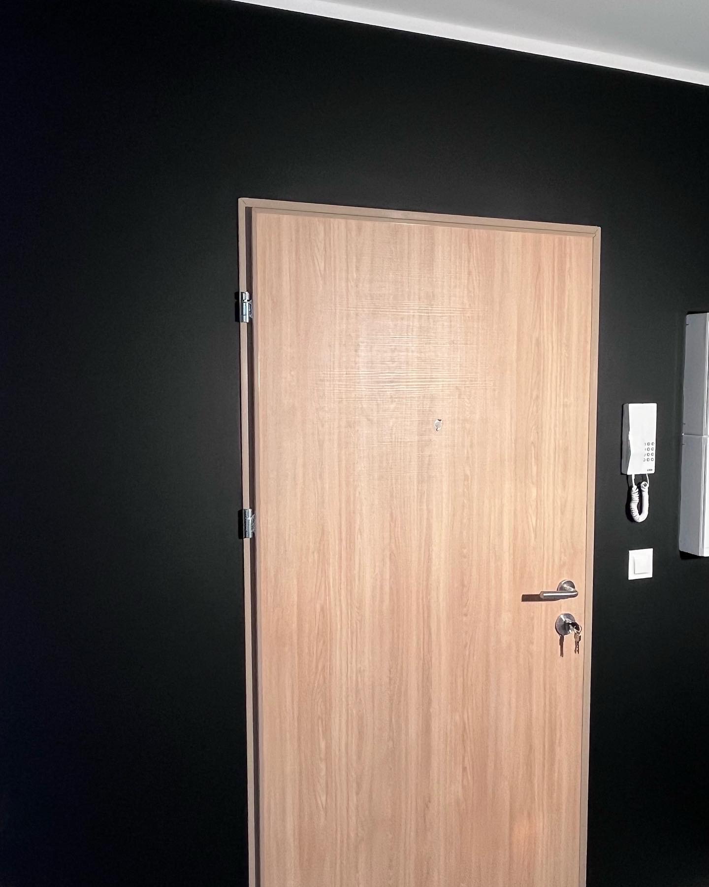 Jasne, drewnopodobne drzwi wejściowe z wizjerem i kluczami w zamku, na tle czarnej, matowej ściany i domofonu