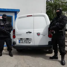 Dwóch umundurowanych ochroniarzy w kominiarkach, stojących obok białego samochodu dostawczego z napisem Ochrona JAG Partner.