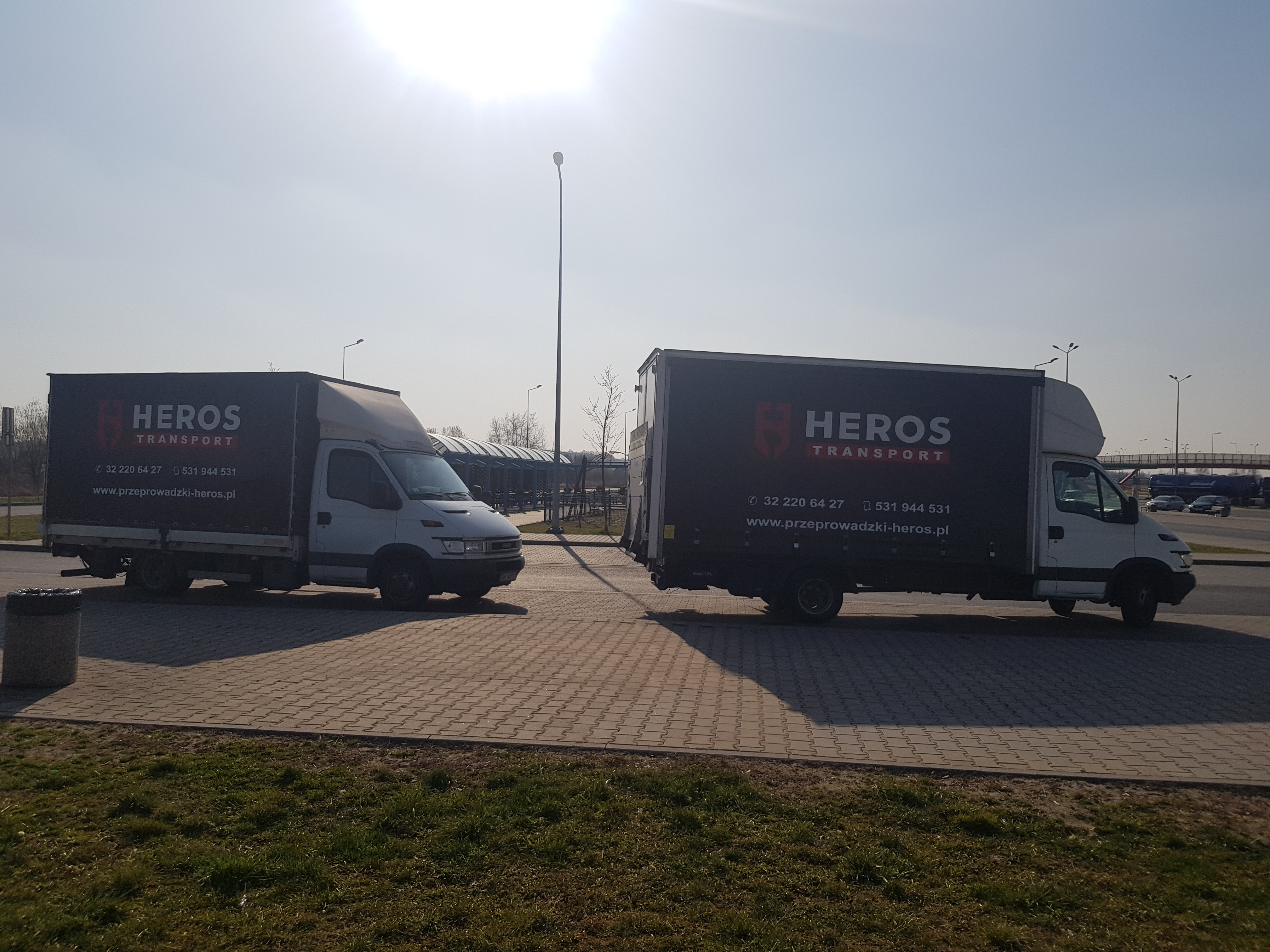 Dwa białe samochody dostawcze z czarnymi plandekami z logo firmy transportowej Heros Transport zaparkowane na brukowanym parkingu w słoneczny dzień.