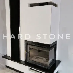 Nowoczesny kominek z obudową z białego materiału i elementami z czarnego kamienia, widoczne niedokończone wykończenia ściany.