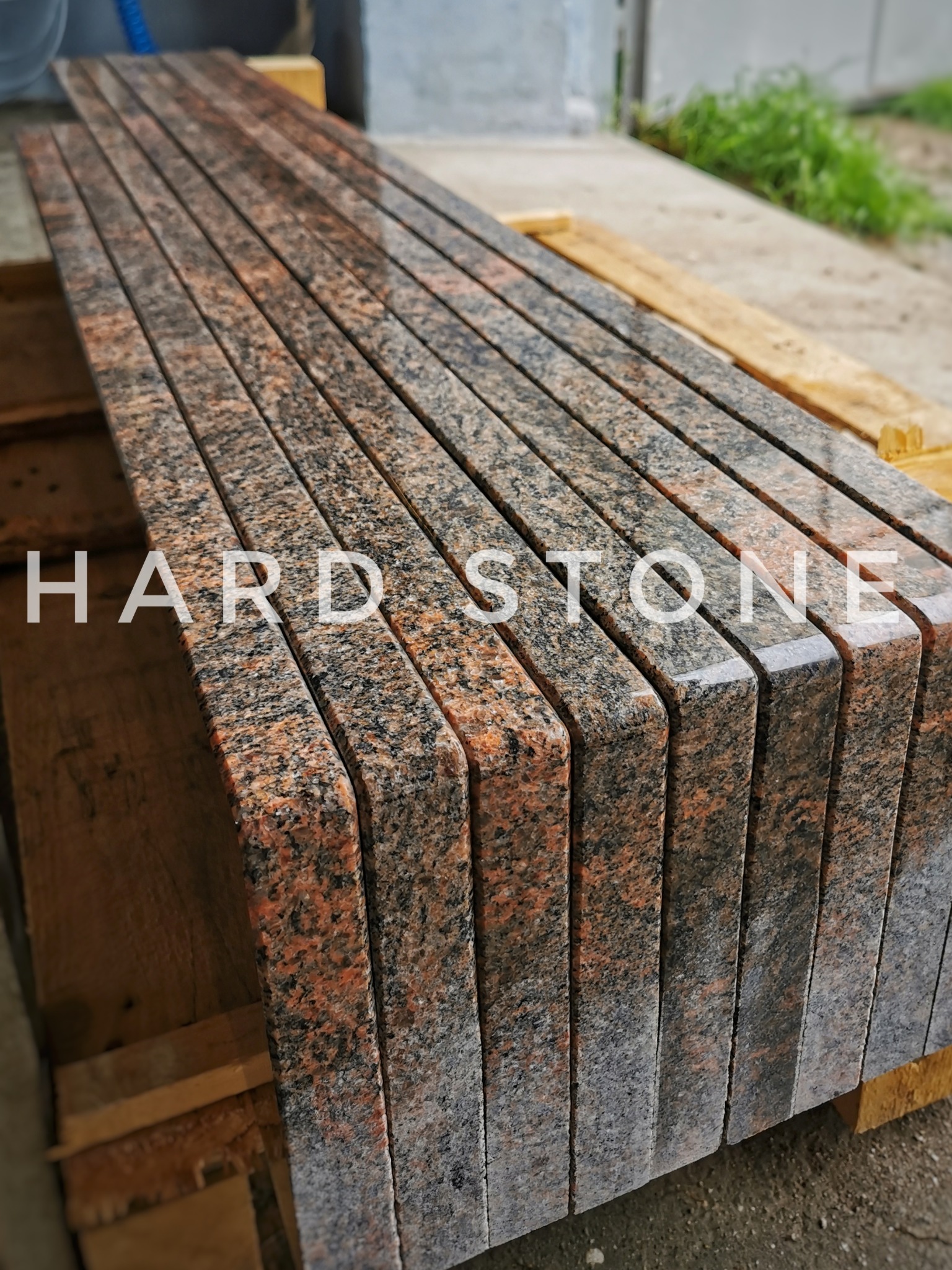 Stos polerowanych blatów kamiennych z granitu w odcieniach czerwieni i czerni, z zaokrąglonymi krawędziami, ułożonych na drewnianej palecie, z widocznym logo 'HARD STONE' na pierwszym blacie.