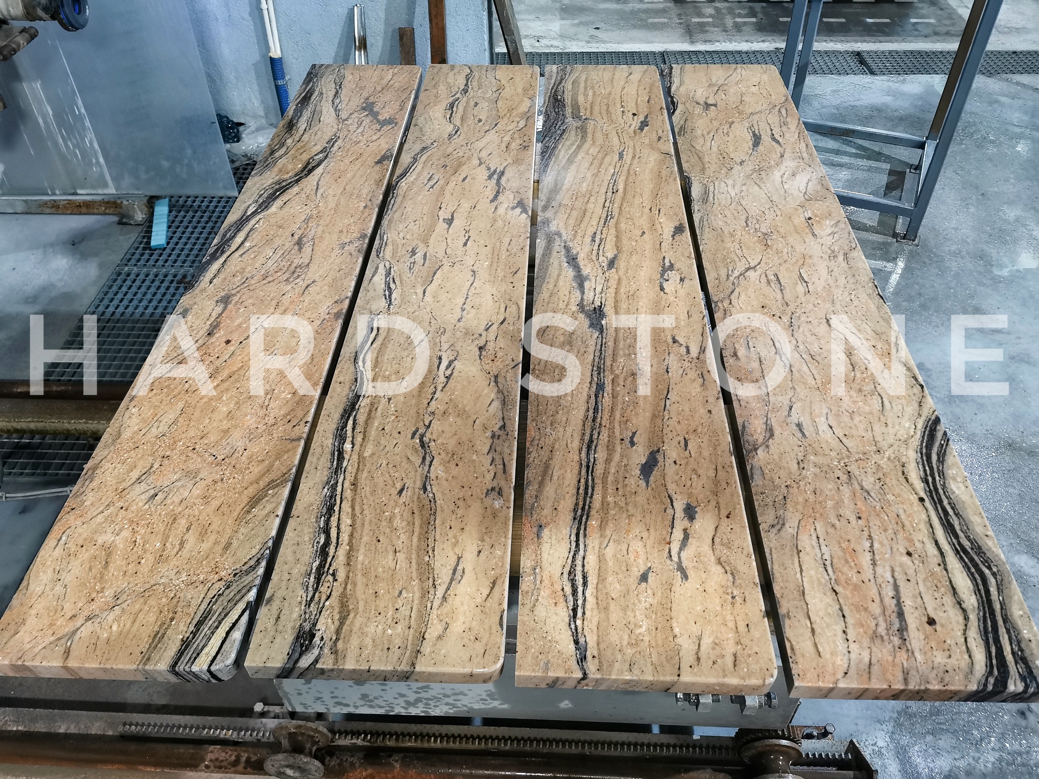 Cztery płyty z jasnego granitu z ciemnymi żyłkami ułożone obok siebie na metalowym stole roboczym w warsztacie kamieniarskim.