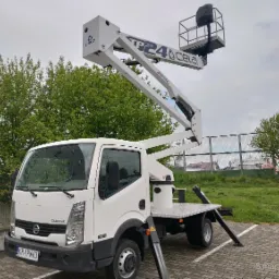 Biały podnośnik koszowy CELA TP24 na samochodzie Nissan Cabstar 35.11, zaparkowany na kostce brukowej, z wysuniętym ramieniem i koszem w tle zieleni drzew i ogrodzenia.
