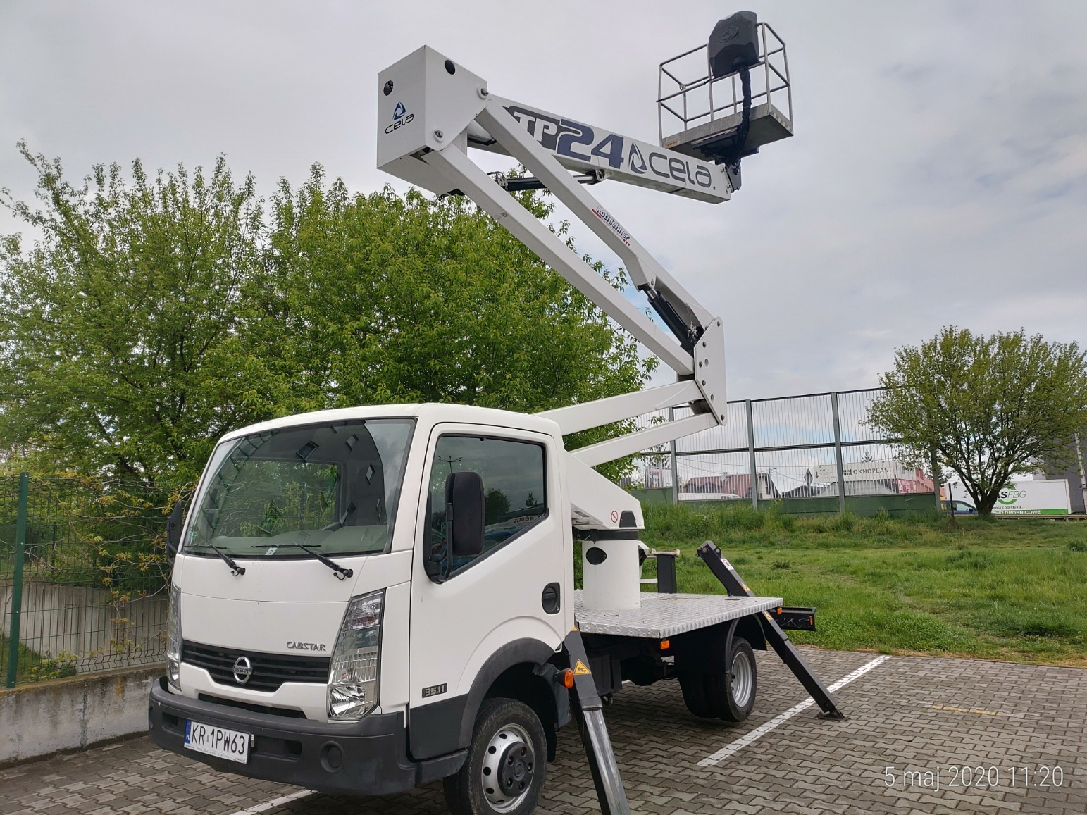 Biały podnośnik koszowy CELA TP24 na samochodzie Nissan Cabstar 35.11, zaparkowany na kostce brukowej, z wysuniętym ramieniem i koszem w tle zieleni drzew i ogrodzenia.