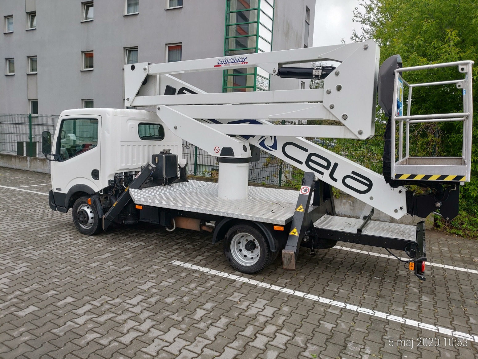 Biały podnośnik koszowy marki Cela zamontowany na podwoziu samochodu ciężarowego, zaparkowany na kostce brukowej przed budynkiem mieszkalnym z zielonymi krzewami w tle.