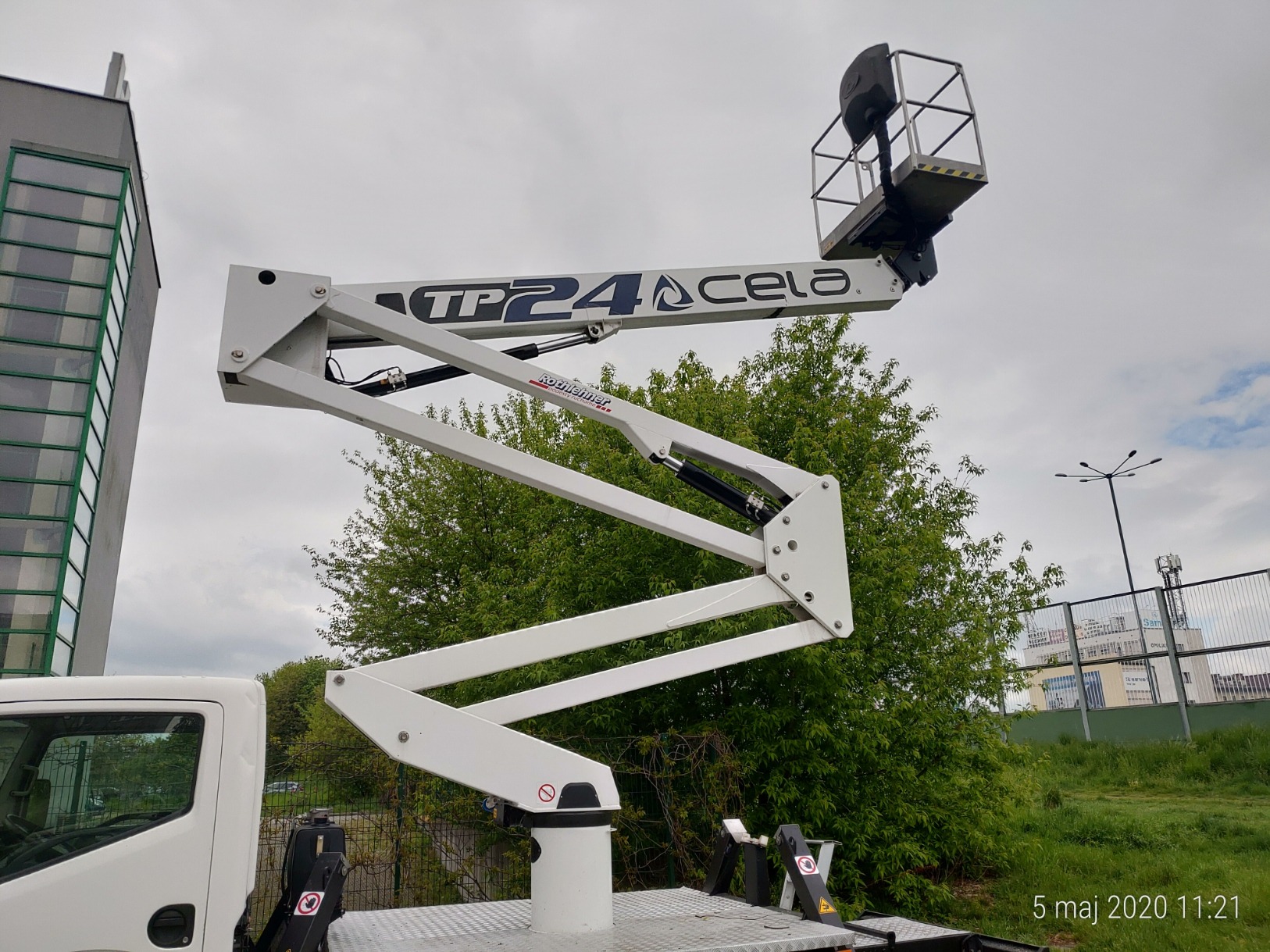 Biały podnośnik koszowy TP24 Cela z wysięgnikiem wzniesionym na tle zieleni i budynku, widok z dołu na platformę roboczą.