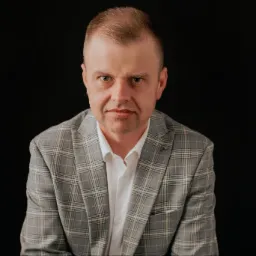 Portret mężczyzny w szarej marynarce w kratę i białej koszuli, na czarnym tle, o intensywnym spojrzeniu w obiektyw.