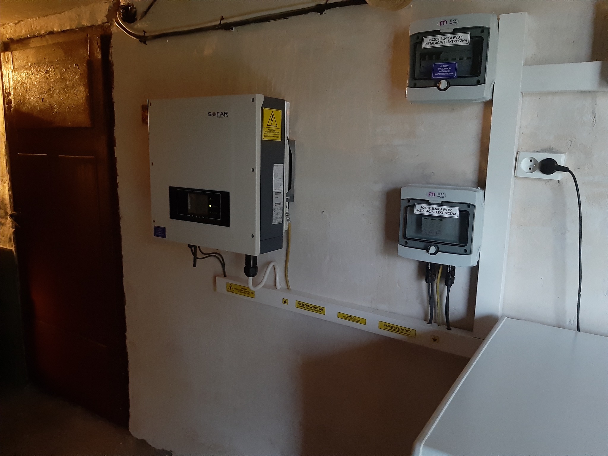 Instalacja fotowoltaiczna w piwnicy z inwerterem Sofar i rozdzielnicami ETI PV AC/DC, z widocznymi naklejkami ostrzegawczymi na listwie montażowej.