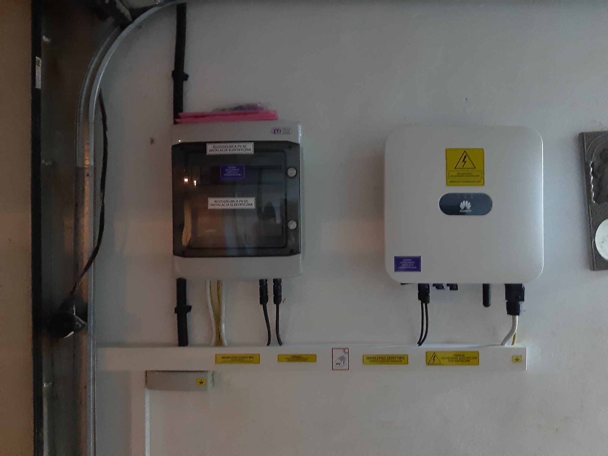 Rozdzielnica PV AC i falownik, zamontowane na białej ścianie wewnątrz pomieszczenia, widoczne kable i oznaczenia ostrzegawcze.