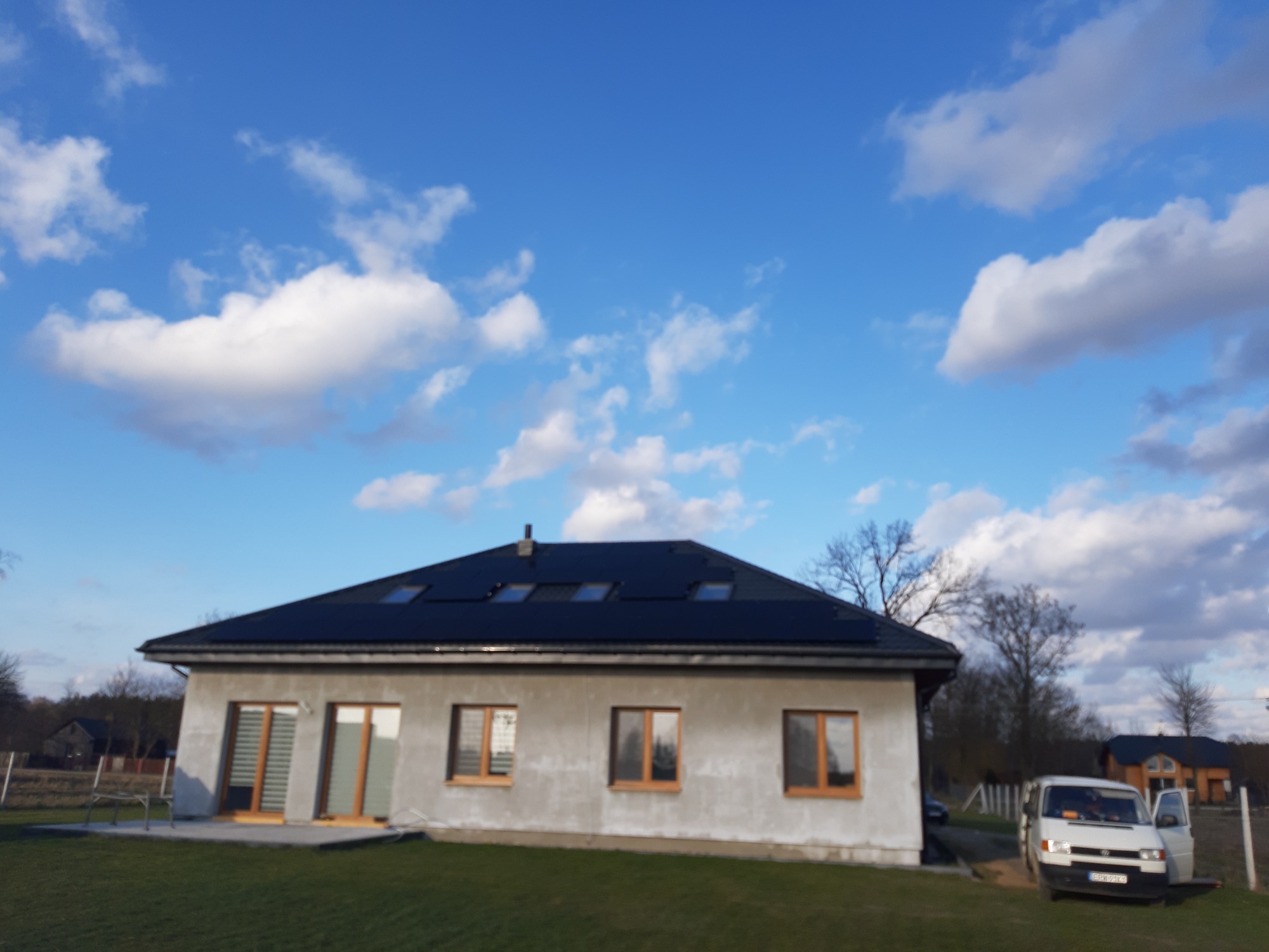 Dom jednorodzinny z zainstalowanymi panelami słonecznymi na dachu, widoczny biały samochód dostawczy z otwartymi drzwiami po prawej stronie, błękitne niebo z obłokami w tle.