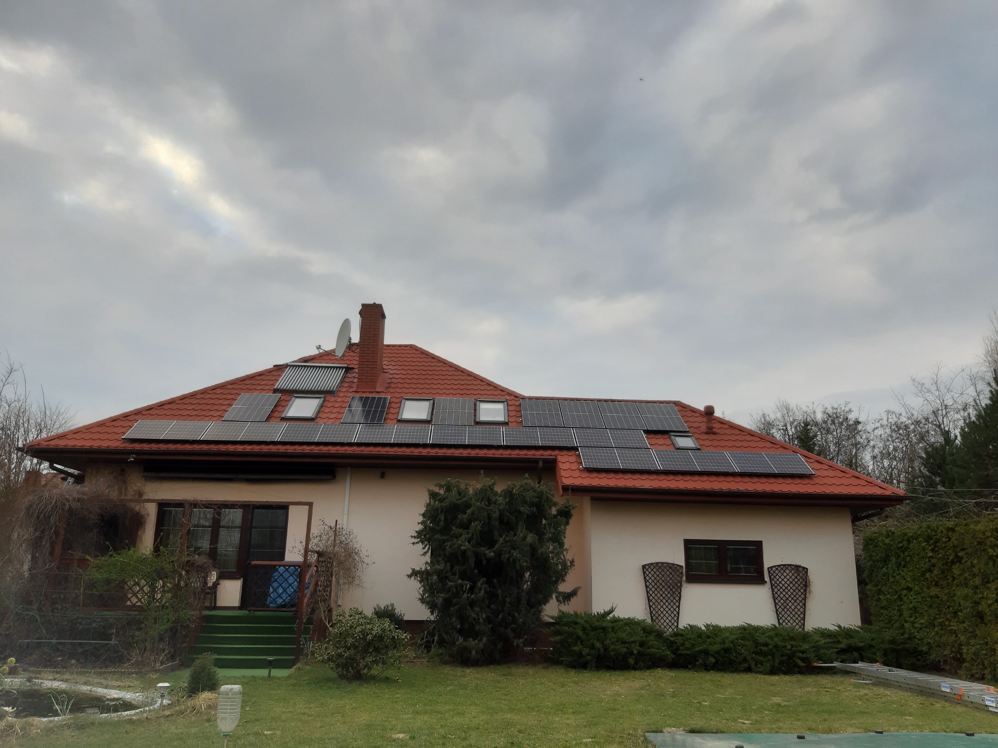 Dom jednorodzinny z czerwonym dachem, na którym zamontowano panele słoneczne i kolektory słoneczne, widoczne na tle pochmurnego nieba.
