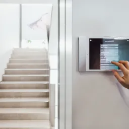 Panel sterowania systemem zarządzania oknami w nowoczesnym domu, zamontowany na ścianie obok schodów, obsługiwany dotykowo przez kobiecą dłoń.