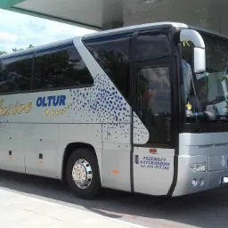 Srebrny autokar turystyczny z napisem 'Exclusive OLTUR' i niebieskimi plamkami, zaparkowany pod zadaszeniem stacji paliw.