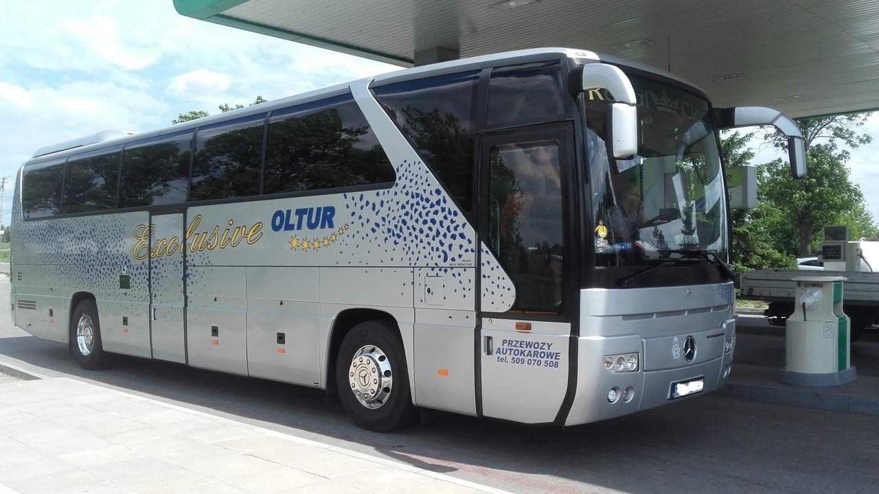 Srebrny autokar turystyczny z napisem 'Exclusive OLTUR' i niebieskimi plamkami, zaparkowany pod zadaszeniem stacji paliw.
