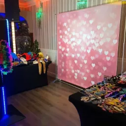 Stanowisko do fotobudki z różowym tłem w serduszka, rekwizytami na stole i oświetleniem LED, ustawione w pomieszczeniu z dekoracyjną tapetą i kryształowym żyrandolem.