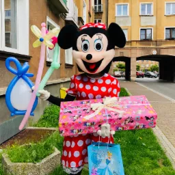 Osoba w kostiumie Myszki Minnie trzyma balonowe dekoracje, prezent w różowym papierze i torbę z wizerunkiem Kopciuszka na tle budynku.
